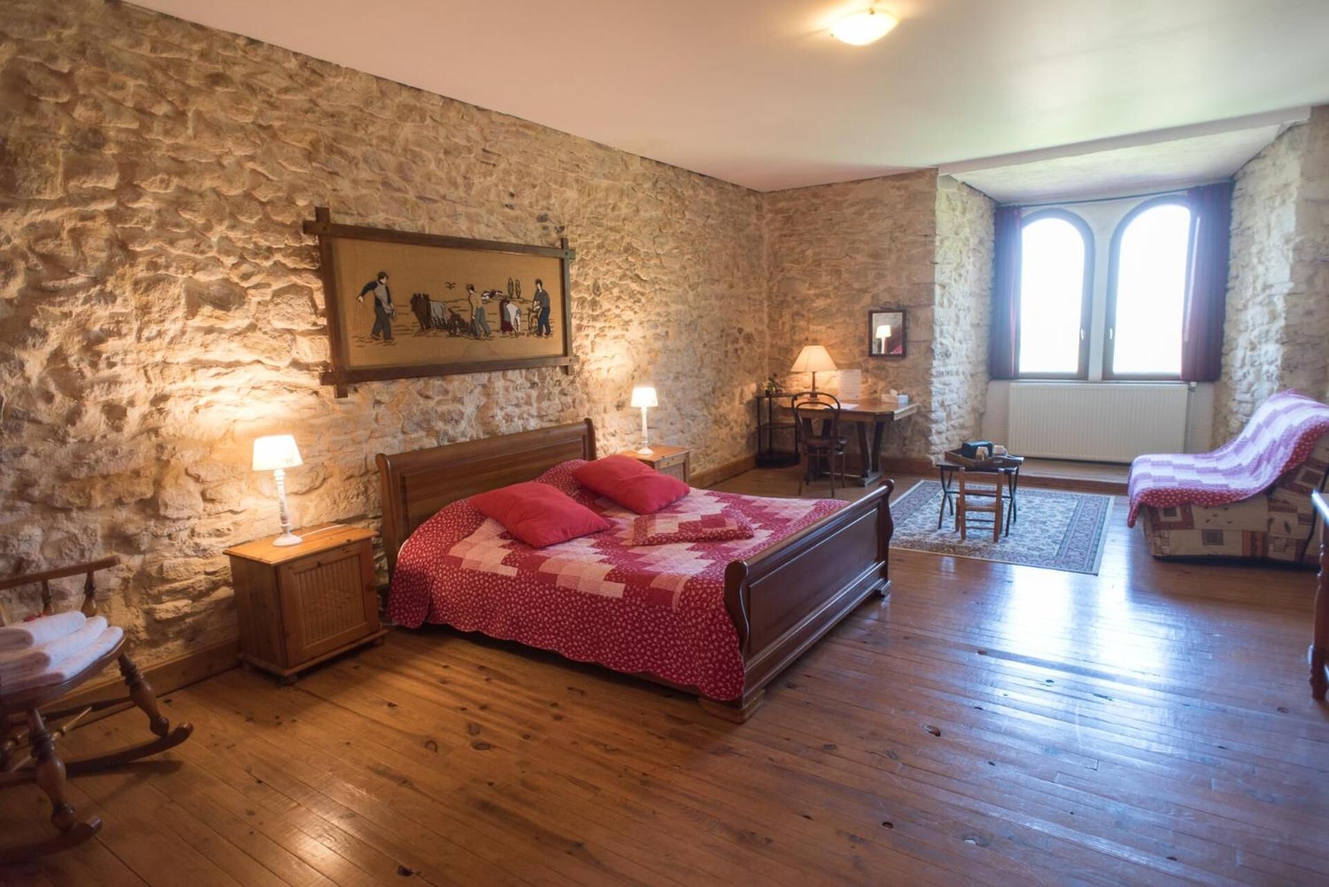 A bedroom with a stone wall, wooden floor, a bed with a red quilt, a rocking chair, a desk, and a chair.