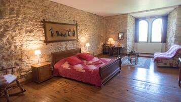 A bedroom with a stone wall, wooden floor, a bed with a red quilt, a rocking chair, a desk, and a chair.