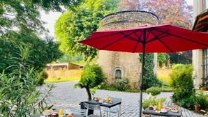 An outdoor patio with tables and chairs, surrounded by greenery and a stone building.