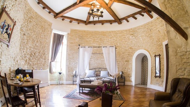 A cozy room with a stone wall, a wooden ceiling, a bed with white curtains, a dining table with chairs, and a leather armchair.
