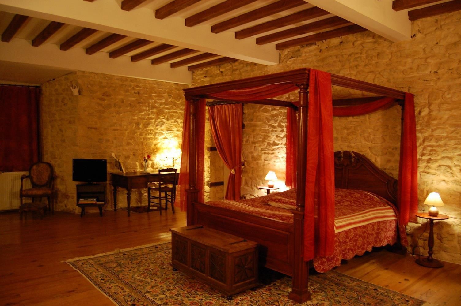 A four-poster bed with red drapes, a wooden desk with a chair, a television, and a lamp on a stone wall.