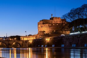 Un castillo histórico con una torre redonda, situado junto a un río con un puente en primer plano.