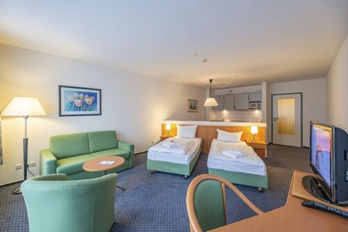 A hotel room with a green sofa, two single beds, a desk with a TV, and a small table.