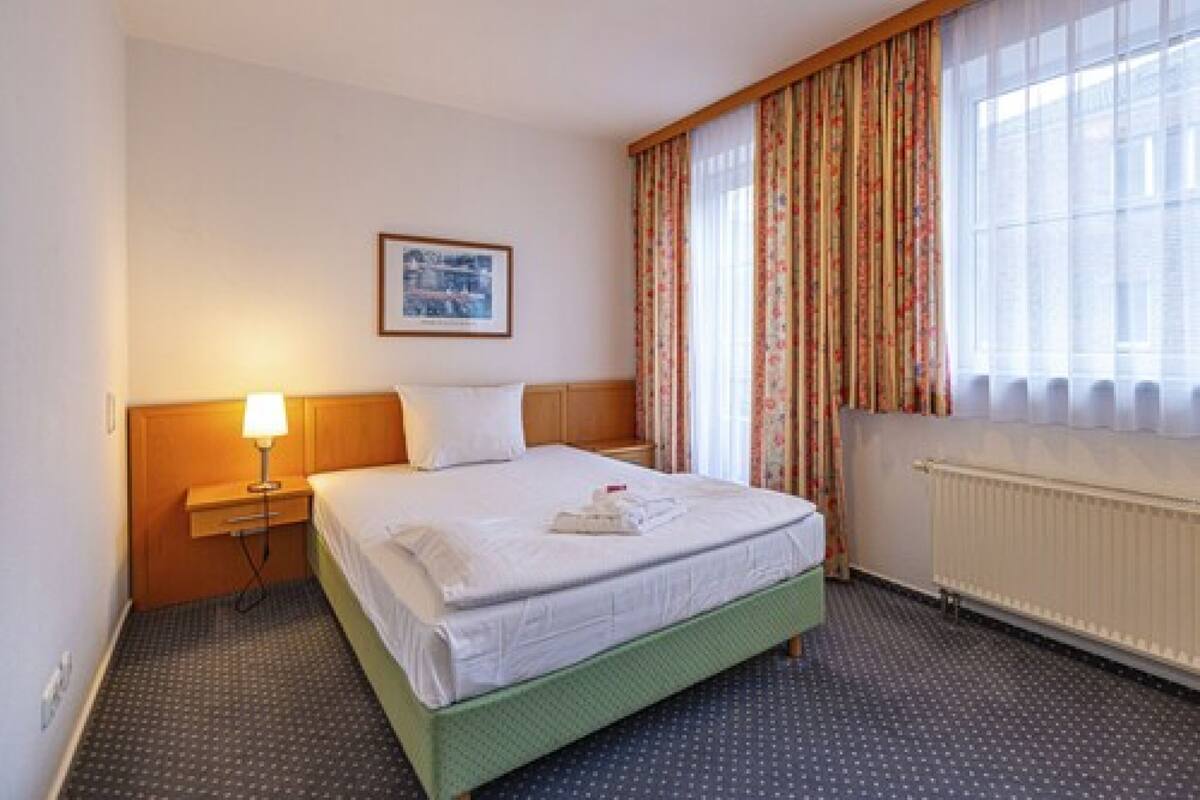 A hotel room with a bed, a nightstand with a lamp, a framed picture on the wall, and a window with curtains.