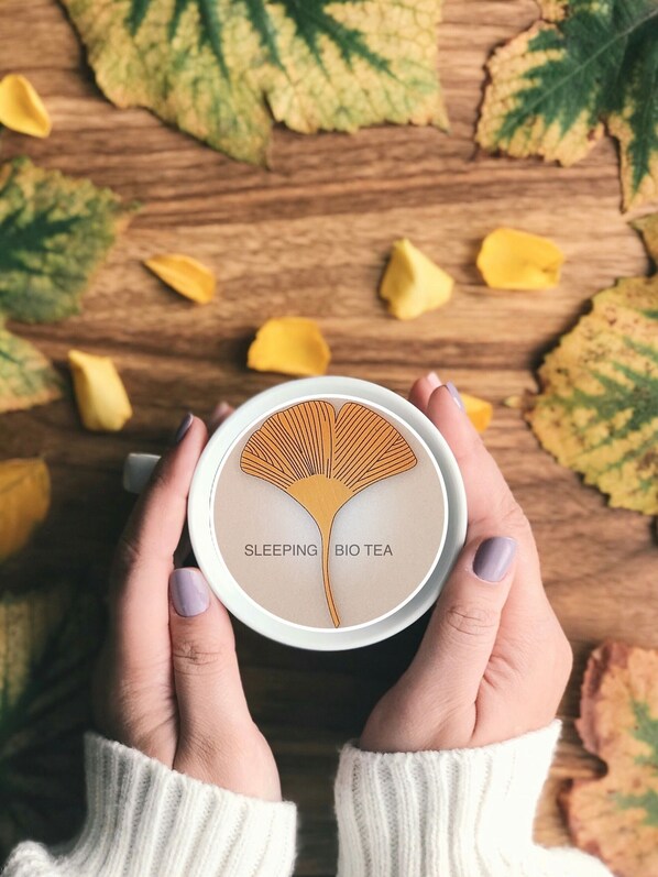 A person holding a container labeled "SLEEPING BIO TEA" with a leaf design, surrounded by autumn leaves on a wooden surface.
