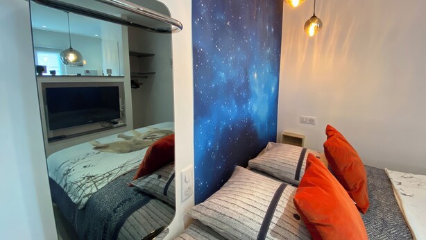 Une chambre à coucher avec un lit, des oreillers, une table de chevet, un miroir, une télévision et une fresque murale représentant un ciel étoilé.