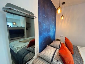 A bedroom with a bed, pillows, a nightstand, a mirror, a TV, and a starry wall mural.