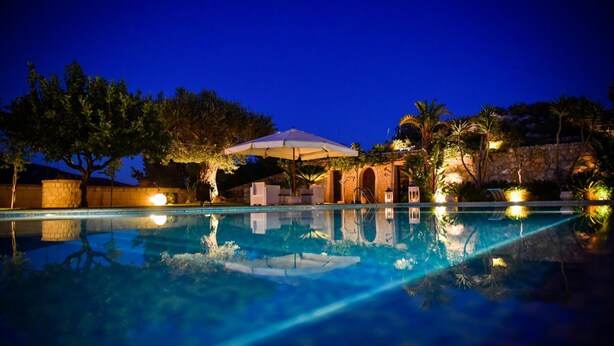 Ein Pool bei Nacht mit klarem Wasser, umgeben von Palmen und einer Steinmauer.