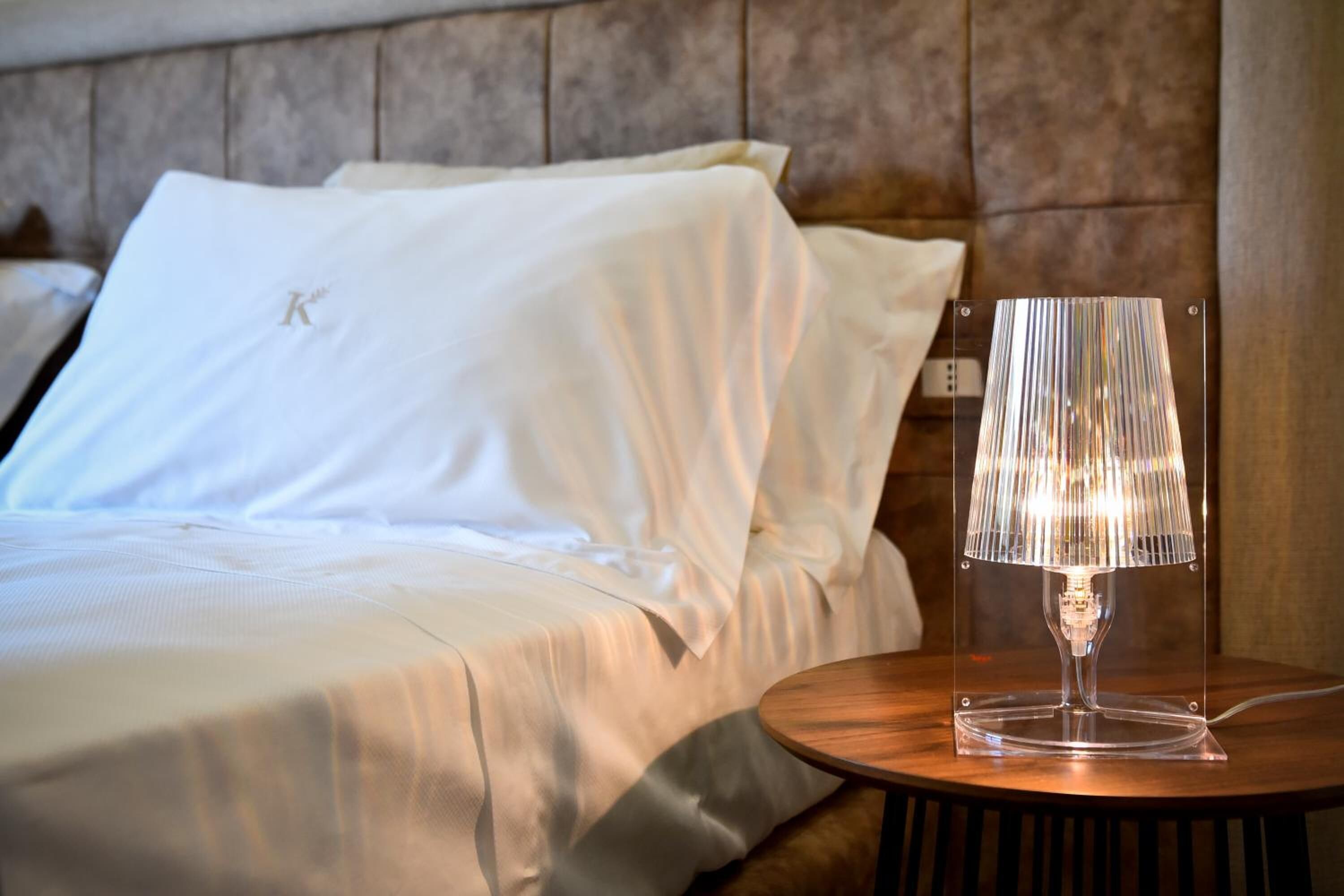 A neatly made bed with white linens and a clear lamp on a wooden bedside table.