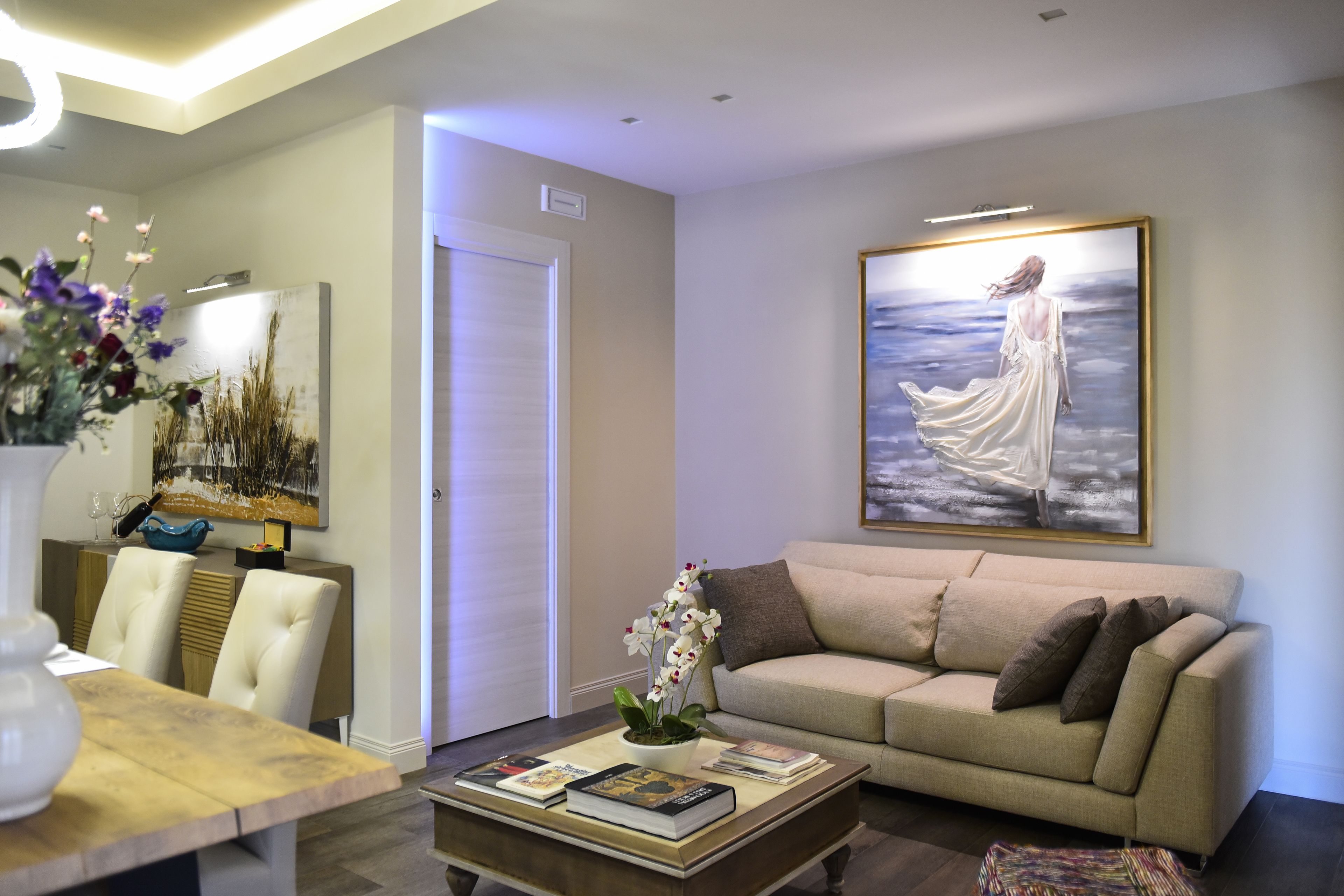 Un salón con un sofá color beige, una mesa de centro con libros, un jarrón con flores y un cuadro en la pared.