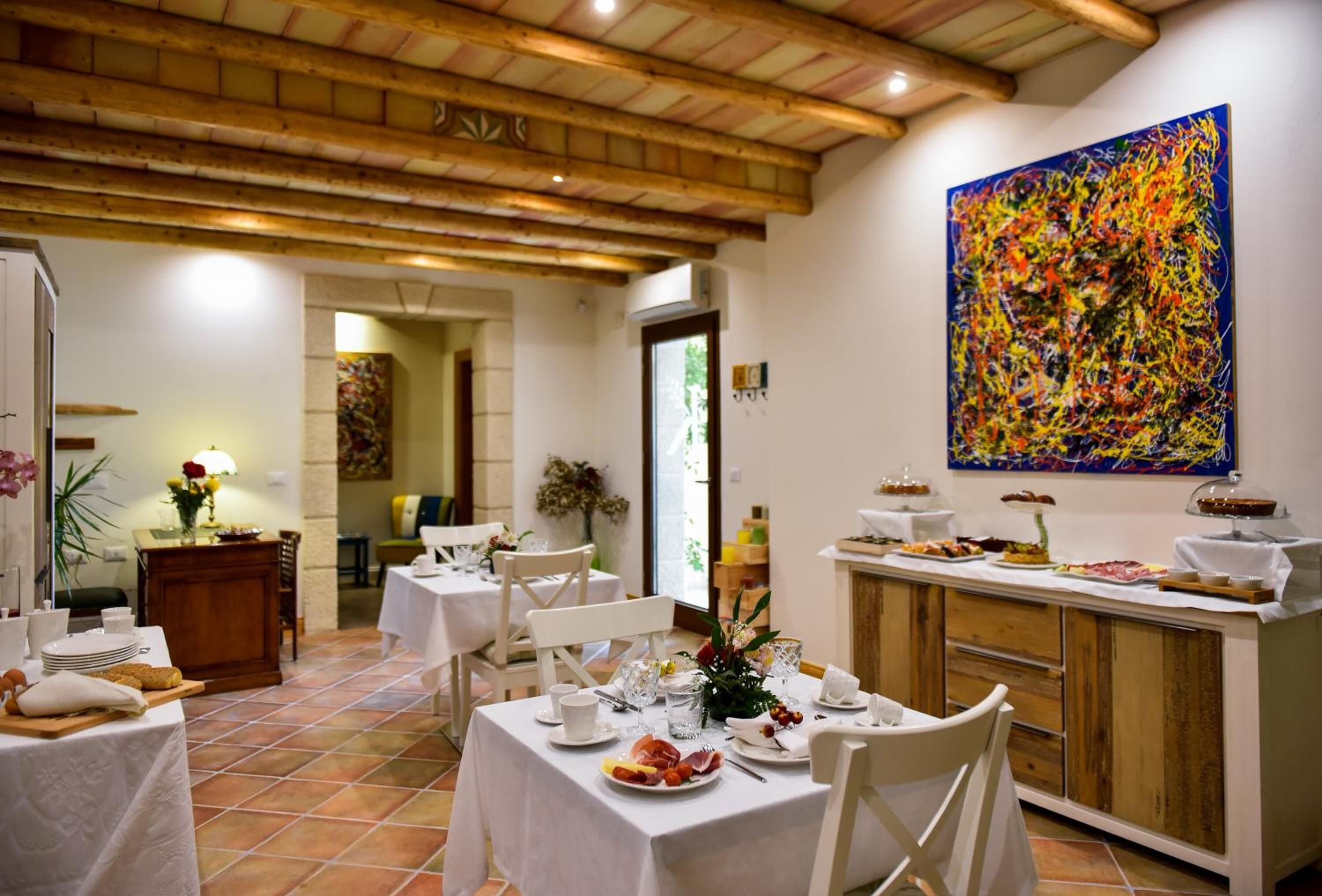 Un coin repas avec des tables préparées pour le petit-déjeuner, une grande peinture abstraite au mur et un plafond en bois.