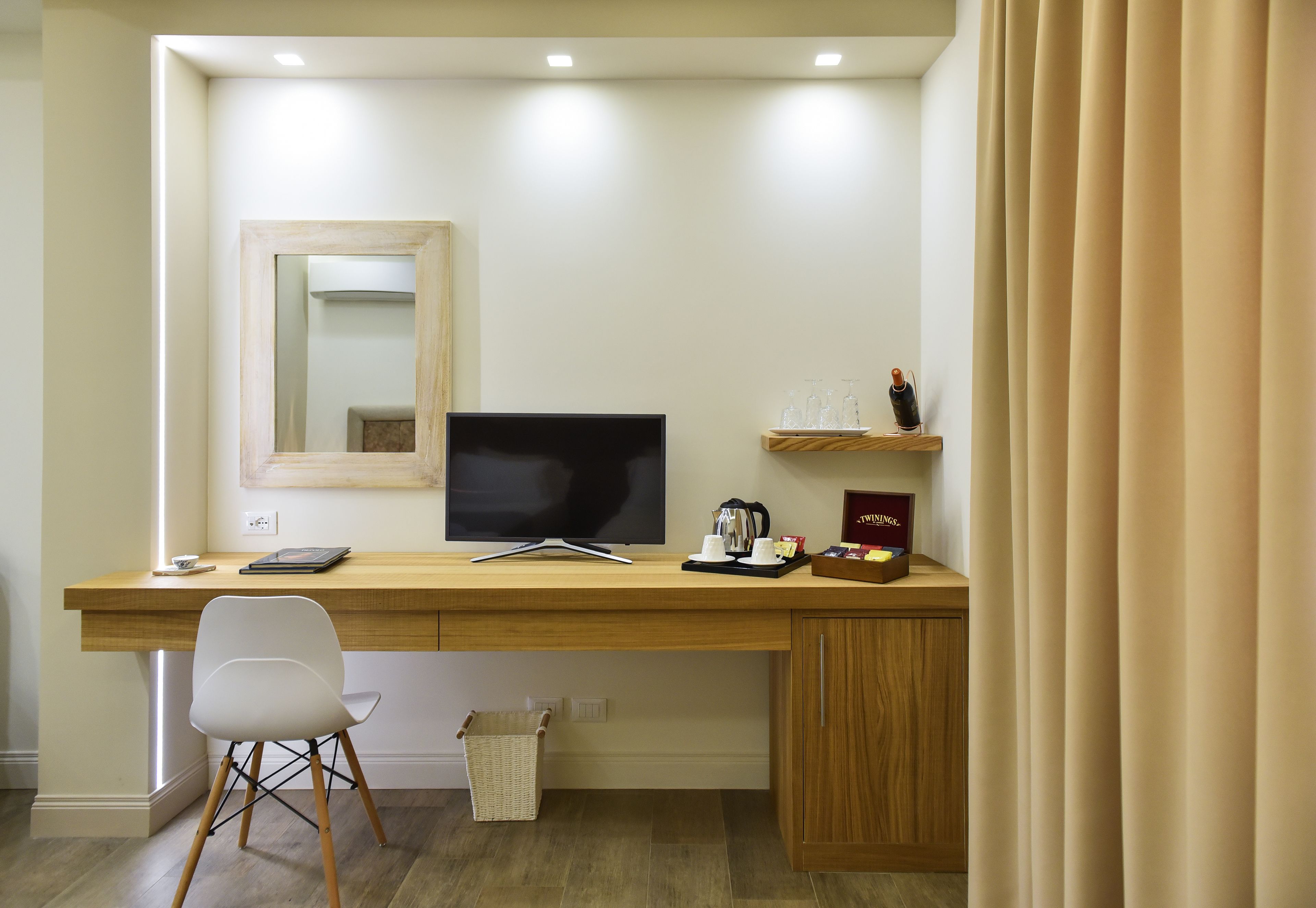 Ein modernes Hotelzimmer mit einem Holztisch, einem Flachbildfernseher, einem weißen Stuhl, einem kleinen Korbwäsches und einem Regal mit einigen Gegenständen.