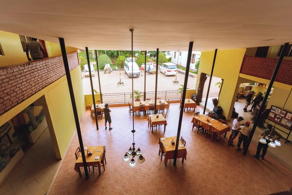 A restaurant with multiple dining tables and chairs, a brick wall on one side, and a view of parked cars outside. - Keys Hotel Mbokomu Branch (Moshi)