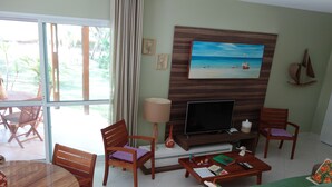 A living room with a TV, wooden furniture, a painting, and a view of the outdoors. - Casa Villa Firenze (Mata de São João)