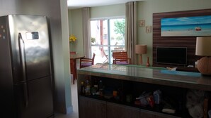 A modern kitchen with a built-in refrigerator, a glass-top dining table, wooden cabinets, and a TV mounted on the wall. - Casa Villa Firenze (Mata de São João)