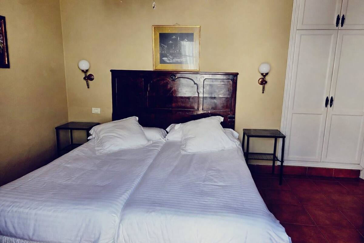 Un lit double avec du linge de lit blanc, dans une chambre dotée d’une tête de lit en bois, de deux tables de chevet, d’une armoire et d’un tableau encadré au mur.