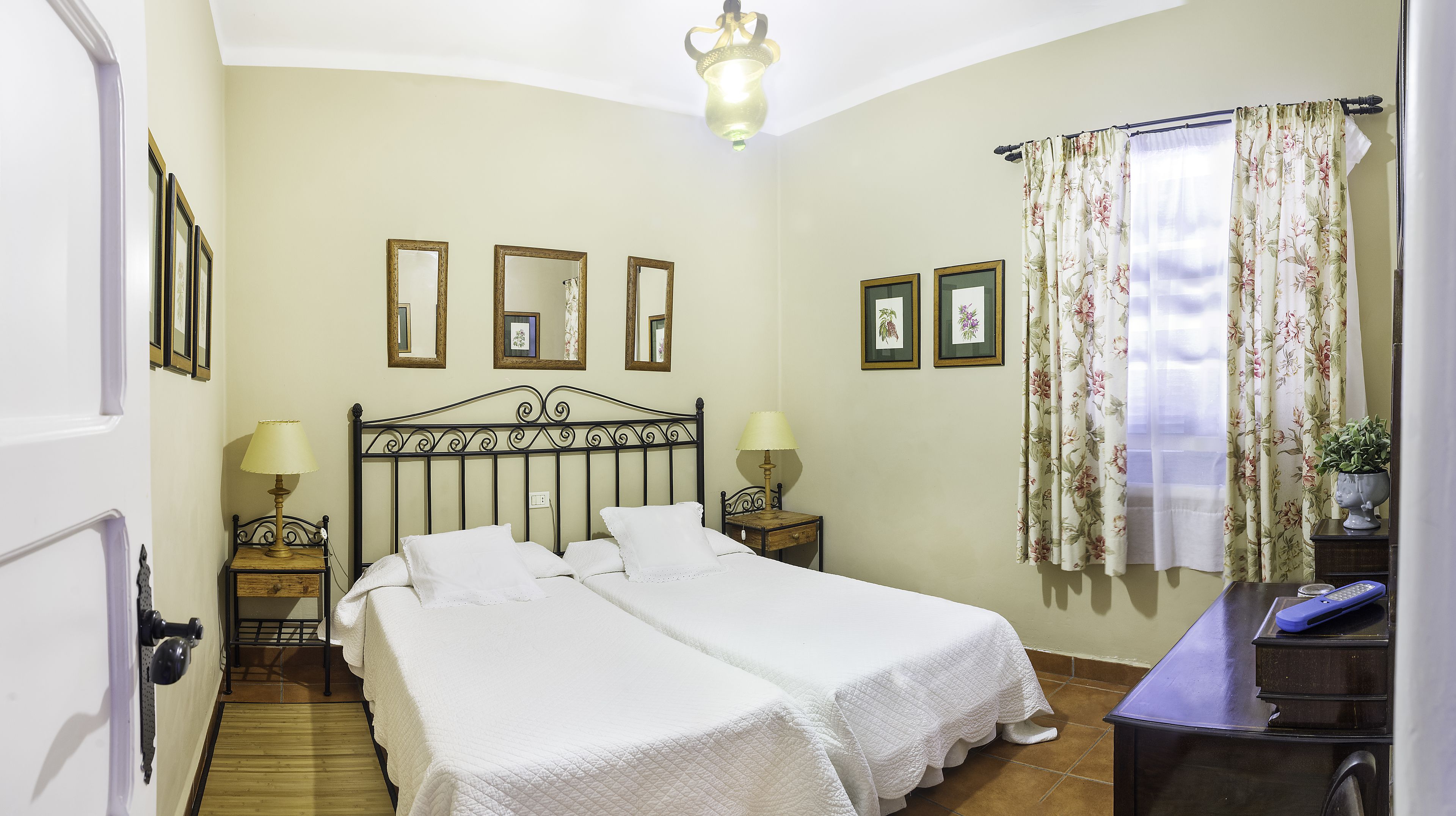 A bedroom with two beds, a wooden floor, a desk with a lamp, and a window with floral curtains.