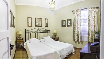 A bedroom with two beds, a wooden floor, a desk with a lamp, and a window with floral curtains.
