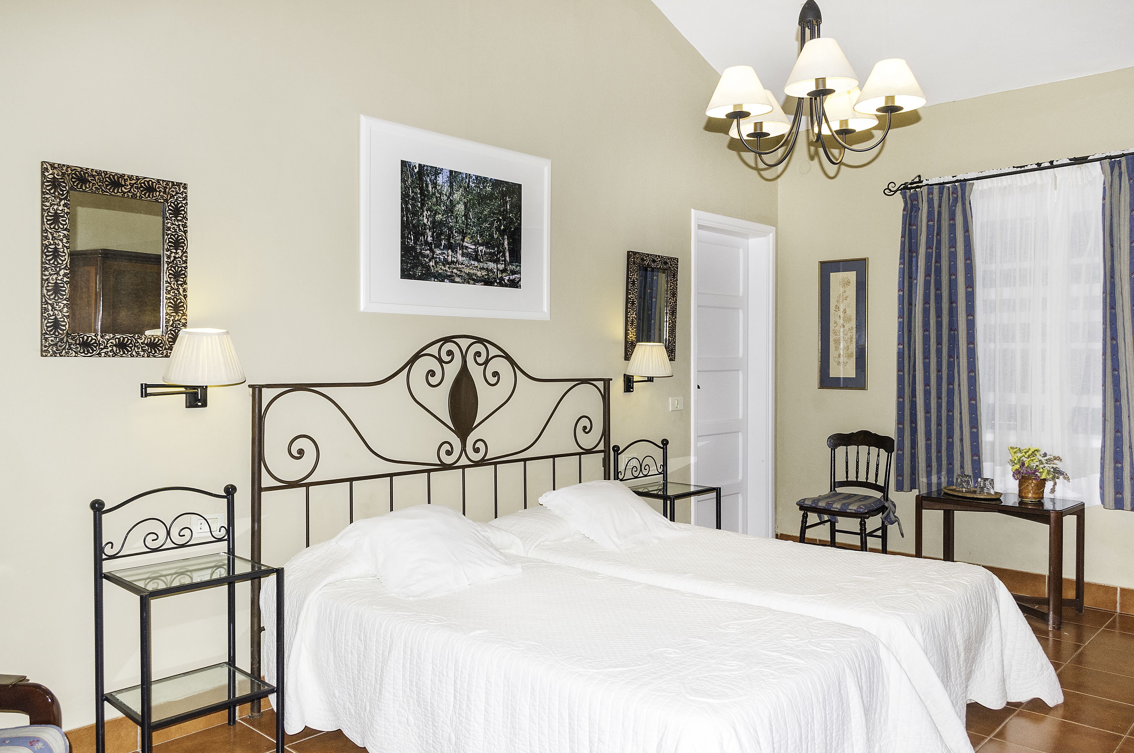 A bedroom with a bed, two bedside tables, a chair, a desk, a chandelier, and framed pictures on the walls.