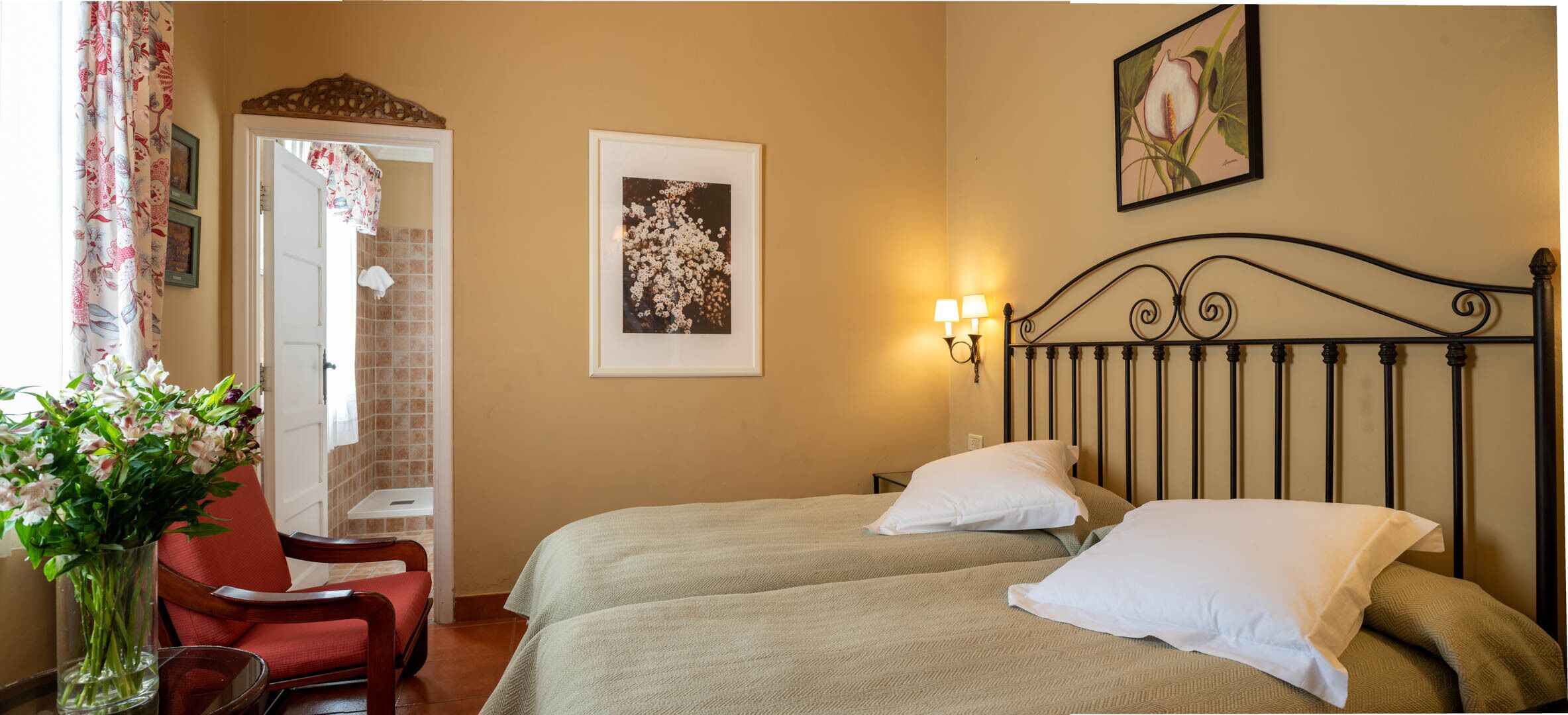 A hotel room with two beds, a red chair, a vase with flowers, and a wall-mounted picture.