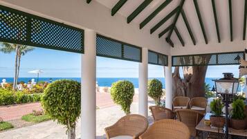 A covered outdoor seating area with wicker furniture, a glass table, and a view of the ocean.