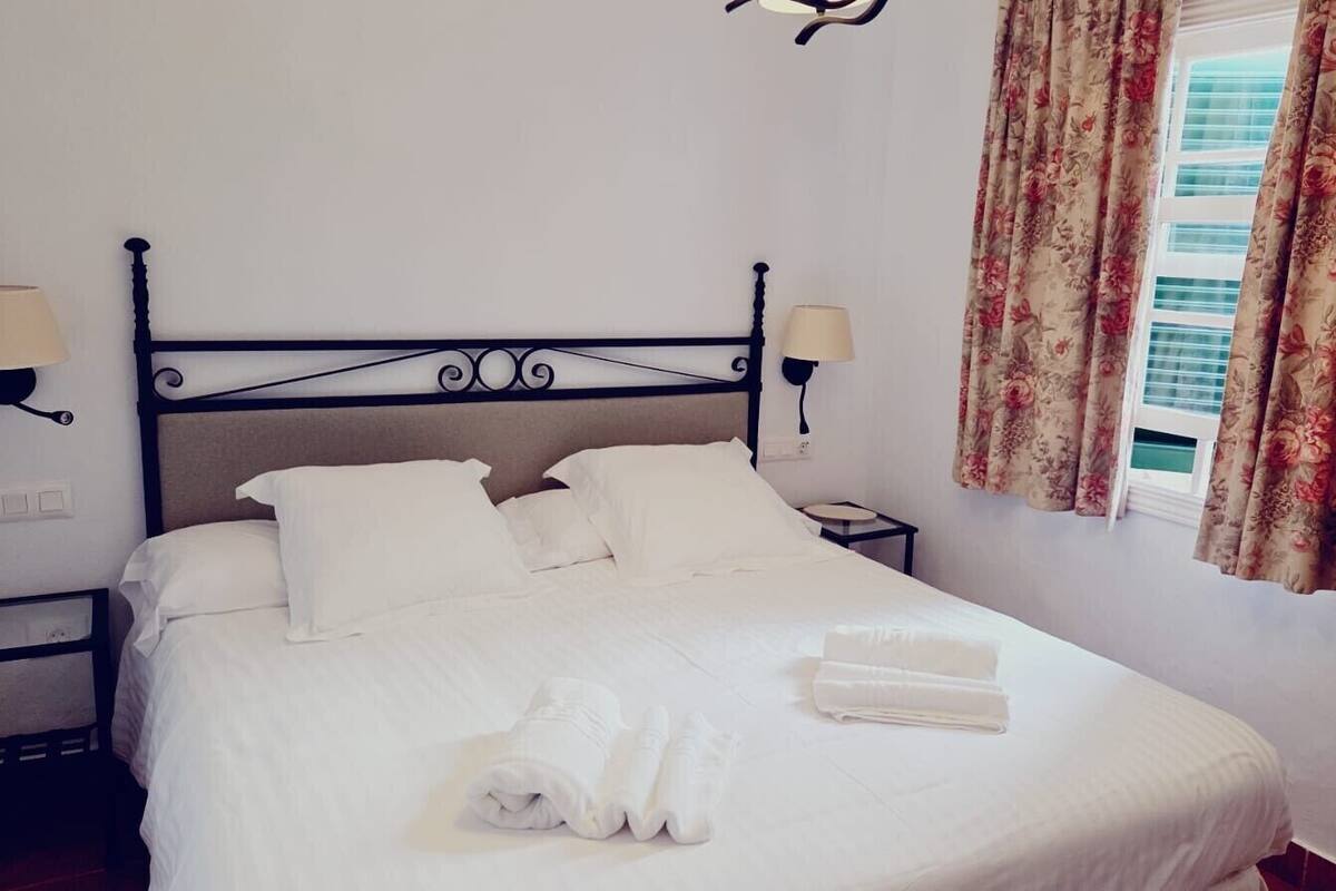 Un lit bien fait, avec du linge de lit et des serviettes blanches, une tête de lit en métal noir, deux lampes de chevet et une fenêtre ornée de rideaux à motifs floraux.
