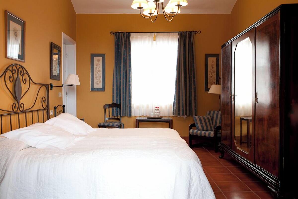 Une chambre d’hôtel avec un grand lit, une armoire, une chaise, une petite table et une fenêtre avec des rideaux.