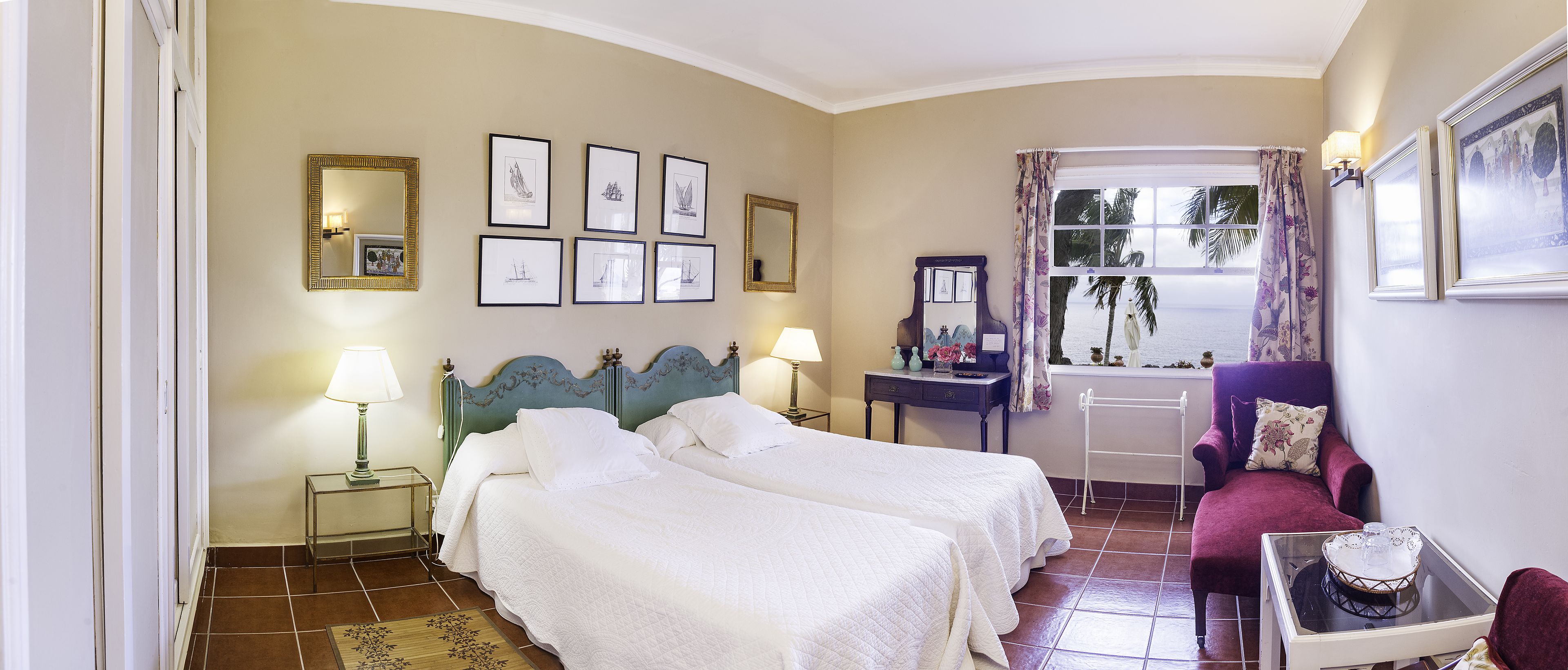 A hotel room with two beds, a vanity with a mirror, a desk with a lamp, and a view of the sea through a window with curtains.