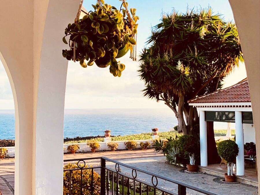 A balcony with a hanging plant, a view of the sea, and a gazebo with potted plants.