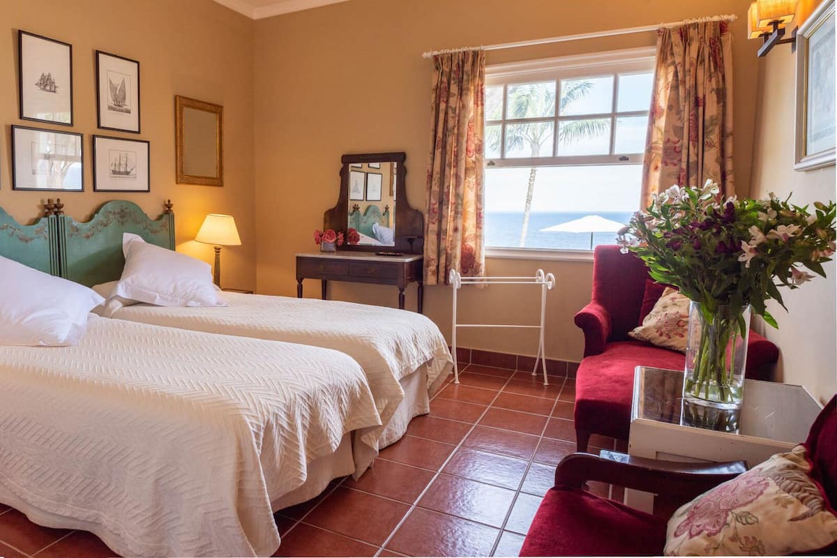 Une chambre d’hôtel avec deux lits, un fauteuil rouge, une petite table sur laquelle est posé un vase de fleurs, et une fenêtre donnant sur la mer.