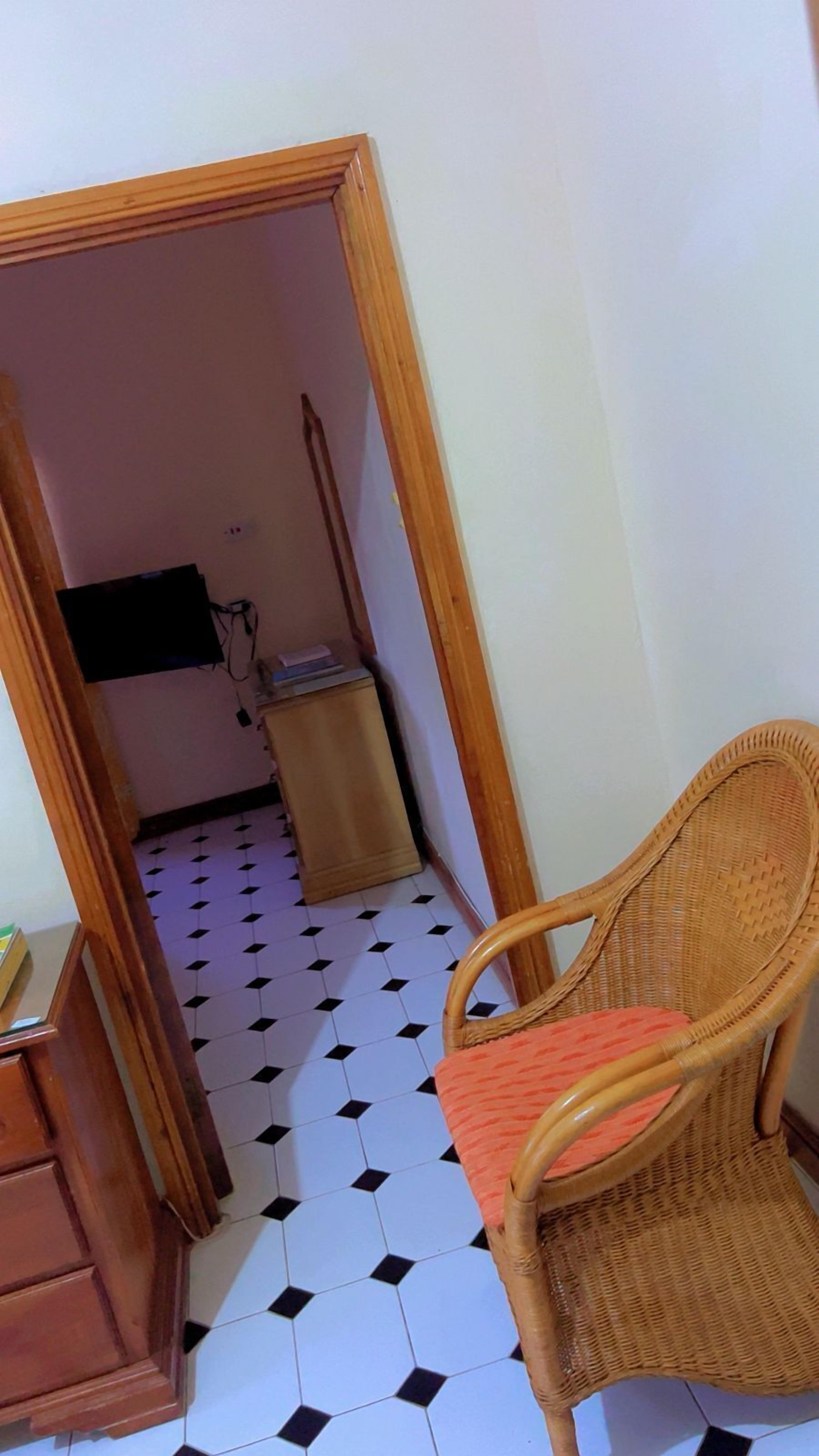 A room with a tiled floor, a wicker chair, a small cabinet, and a television mounted on the wall.