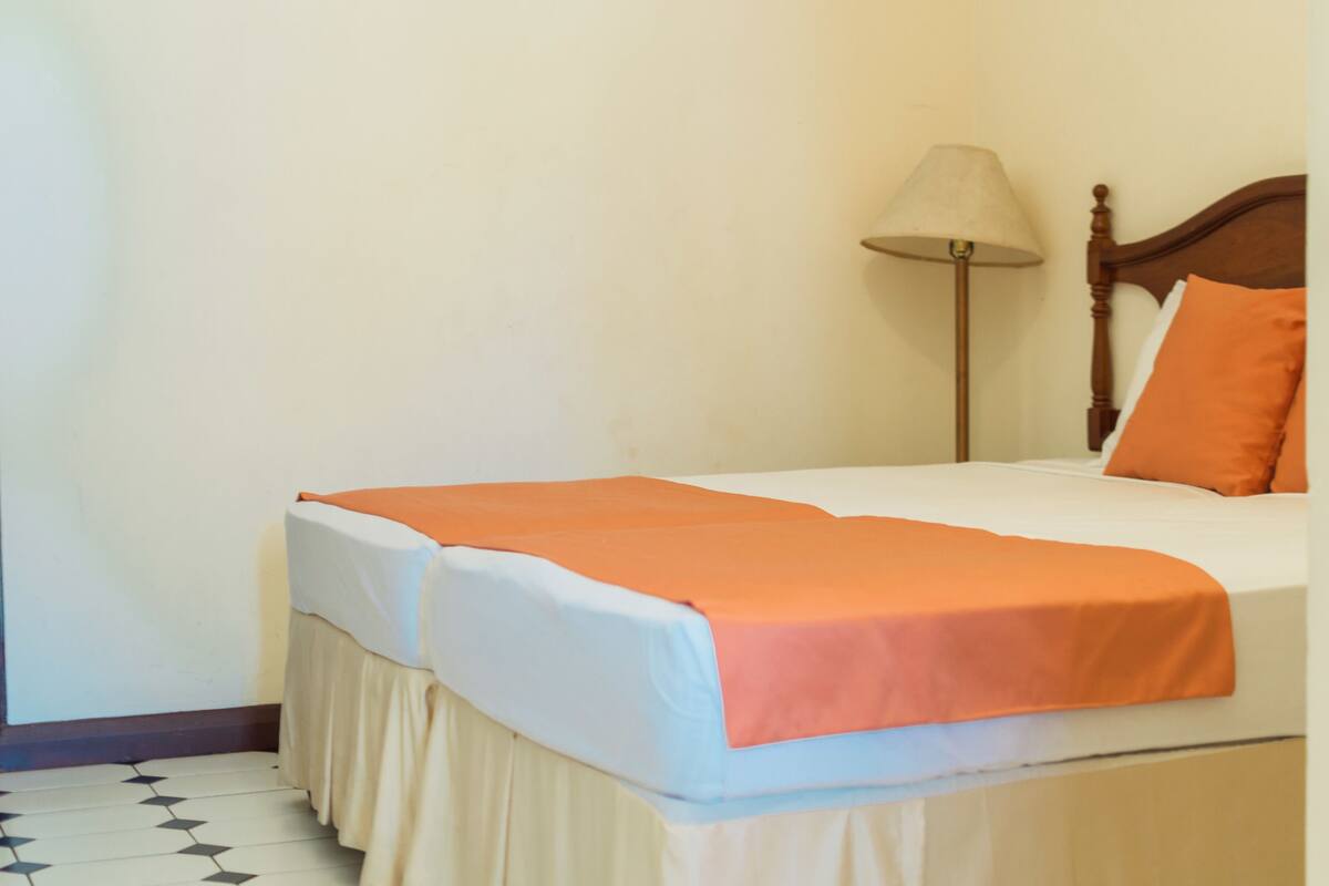 Ein ordentlich bezogenes Bett mit orangefarbener Tagesdecke und weißen Laken, ein Holz-Kopfteil und eine Stehlampe.