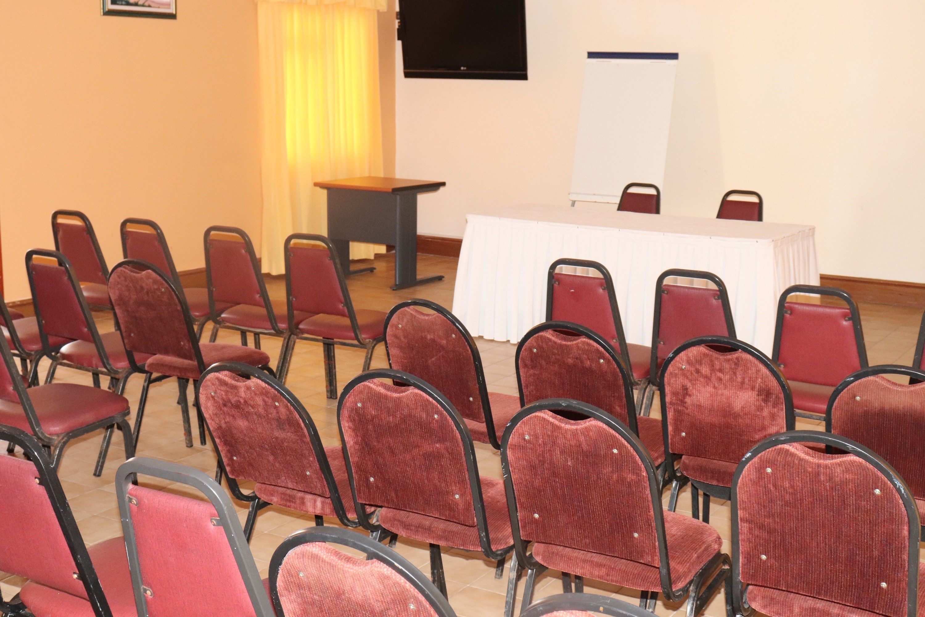 Un salón de conferencias con filas de sillas rojas, una mesa blanca y una pantalla de proyector.
