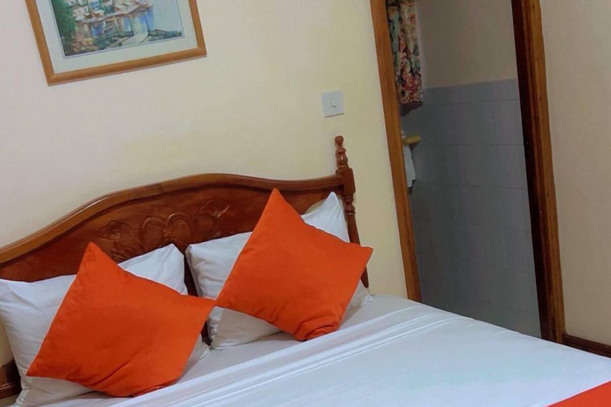 Ein Bett mit weißer Bettwäsche und orangefarbenen Akzenten, ein Holz-Kopfbrett und ein Gemälde an der Wand.