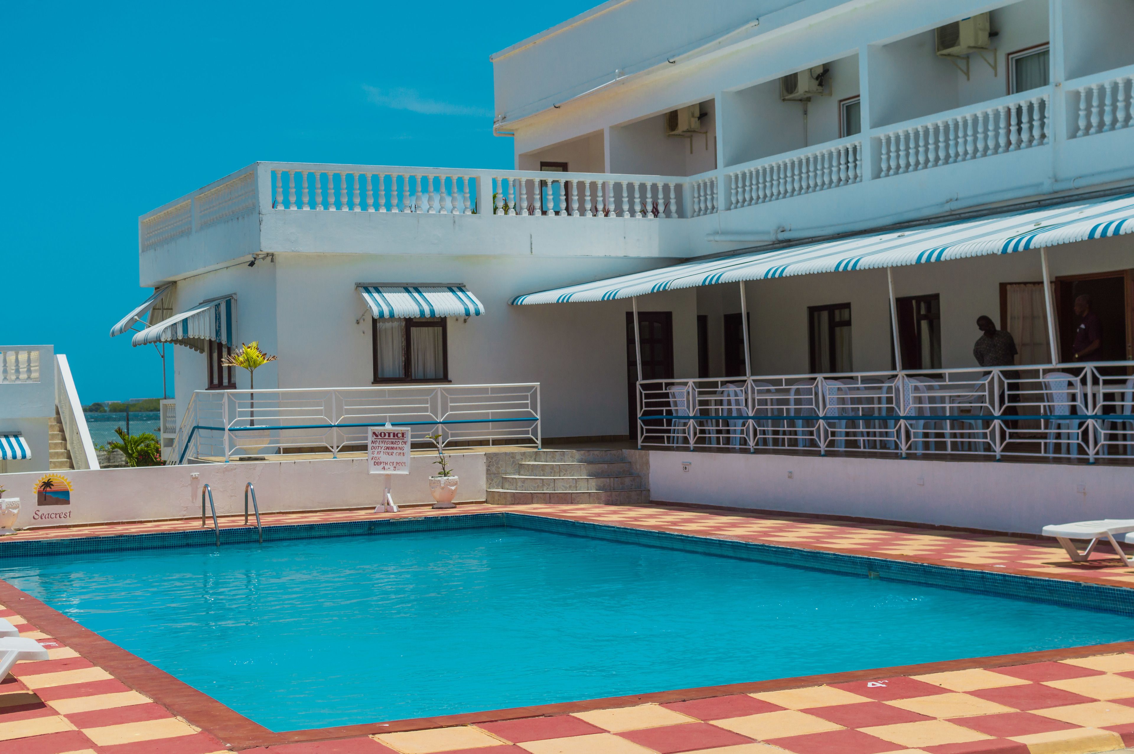 Una piscina con suelo a cuadros, una zona cubierta con sombrillas y un edificio con balcones.