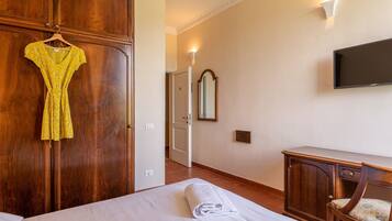 A hotel room with a wooden wardrobe, a bed with a white duvet, a small desk with a chair, and a flat-screen TV mounted on the wall.