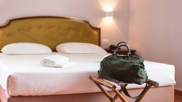 Un letto rifatto con lenzuola bianche, una borsa verde su una sedia di legno e un punto luce a parete.
