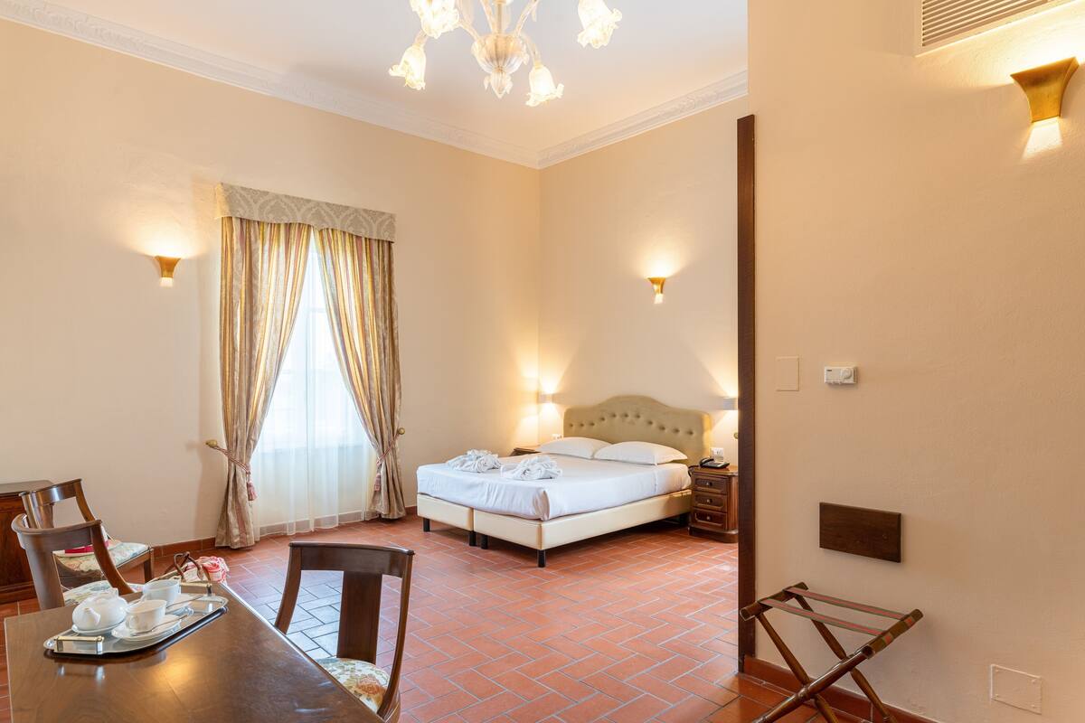 A hotel room with a bed, bedside table, lamp, and a dining area with chairs and a tray with cups and a flower.