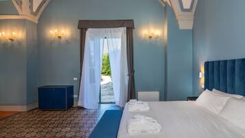 Un dormitorio con una cama grande, un cabecero azul, una mesita de noche y un banco. Se aprecia una ventana con cortinas, un suelo estampado y un banco azul.