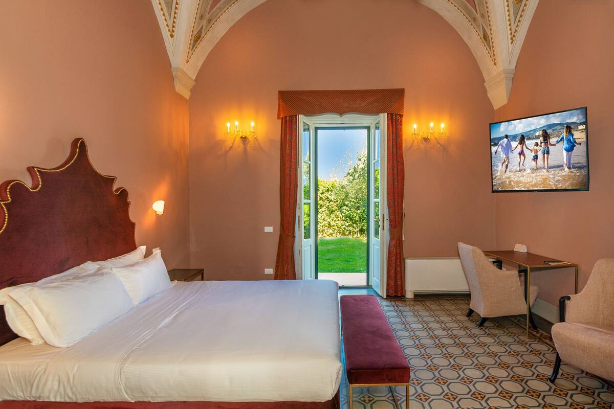 A hotel room with a large bed, a desk, a chair, and a view of a garden through an open door.