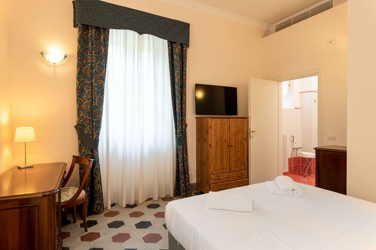 A hotel room with a bed, a wooden wardrobe, a television on a stand, a desk with a chair, and a window with patterned curtains.