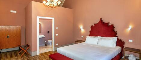 A bedroom with a large bed, a chandelier, a wooden wardrobe, and a bathroom visible through an open door.