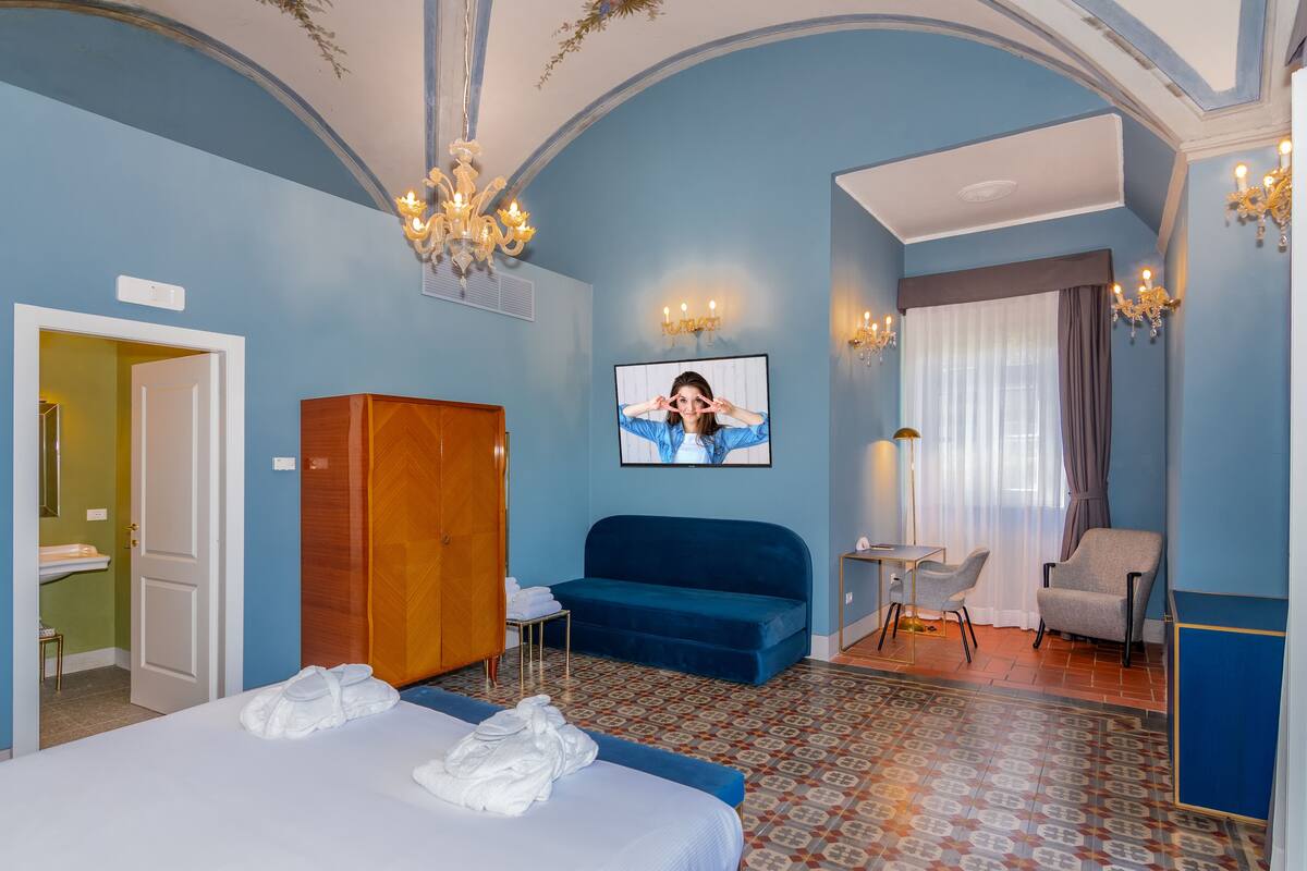 A hotel room with a bed, a blue sofa, a desk with a chair, a wardrobe, and a chandelier.