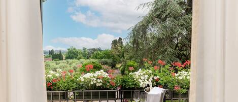 A balcony with a table set for two, overlooking a garden with various flowers and trees.