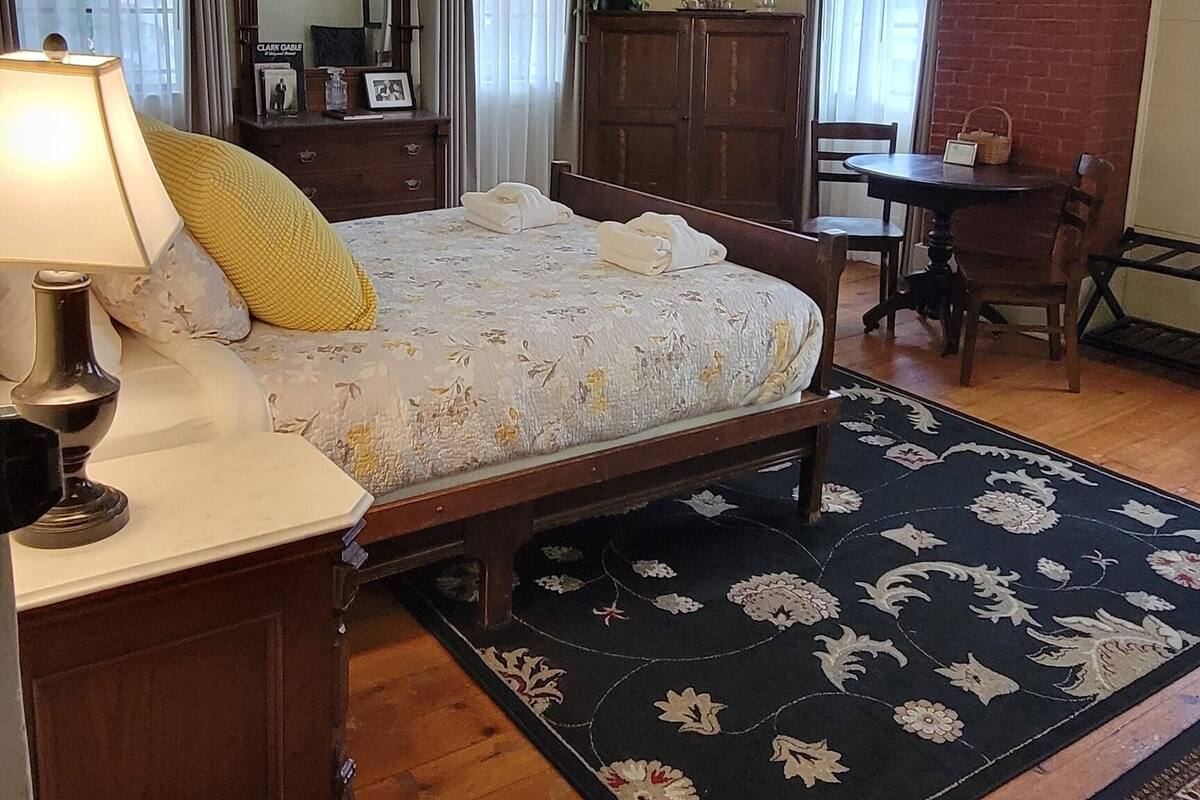 A bedroom with a bed, wooden dresser, and a nightstand with a lamp. There is a mirror above the dresser and a small table with chairs in the corner.