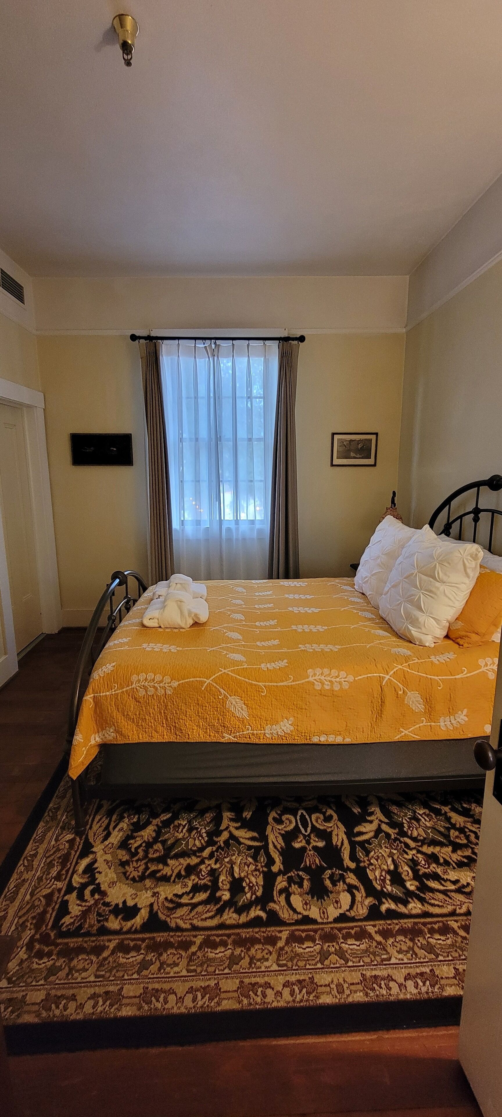 A single bed with a yellow floral bedspread, a black metal frame, and a patterned rug at the foot of the bed.