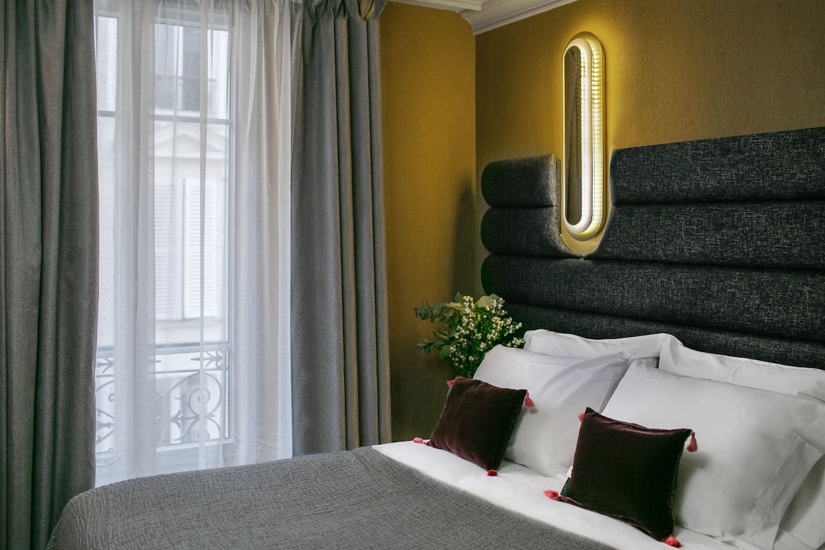 Habitación de hotel con cama, cortinas, una lámpara en la pared y una planta decorativa.