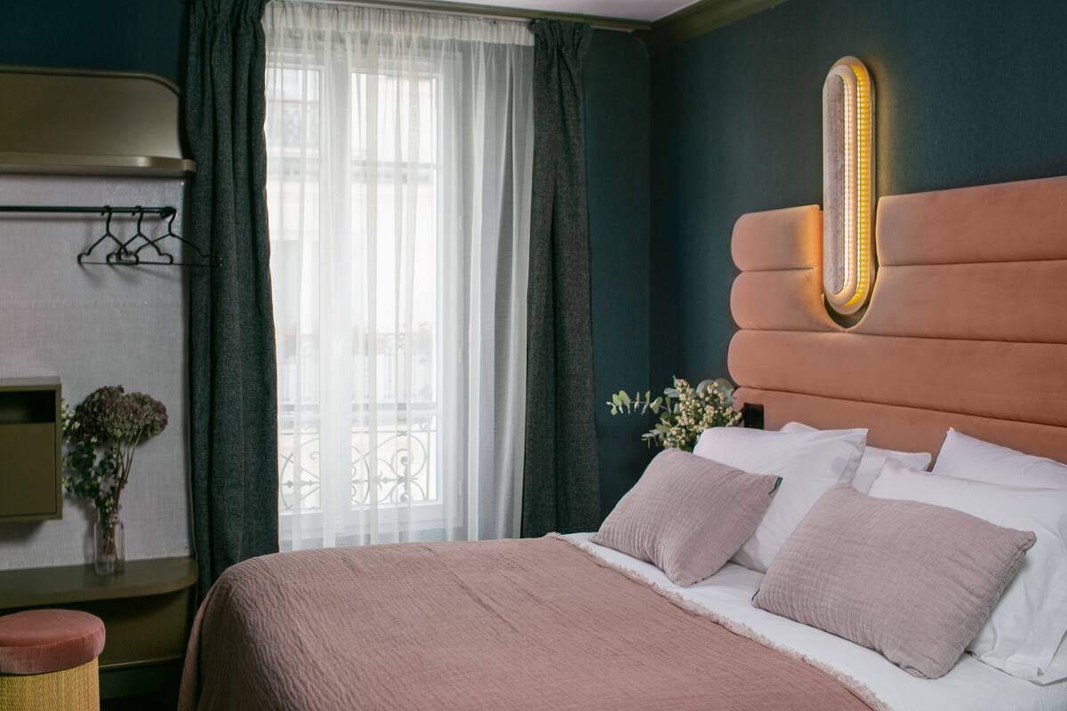 Una cama bien hecha con cabecera que tiene luz integrada. Se ve una ventana con cortinas de gasa y vistas al exterior.
