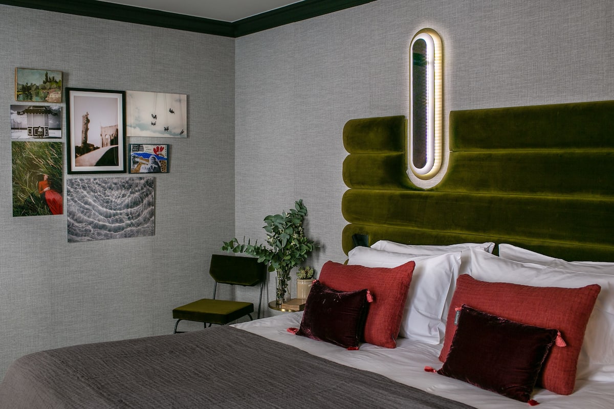 Un dormitorio moderno con un cabecero tapizado en verde, una cama con almohadas blancas y rojas, y una pared decorada con fotografías enmarcadas.