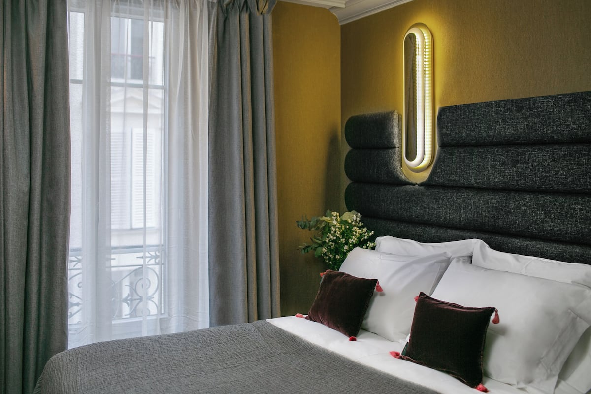Habitación de hotel con cama, cortinas, una lámpara en la pared y una planta decorativa.